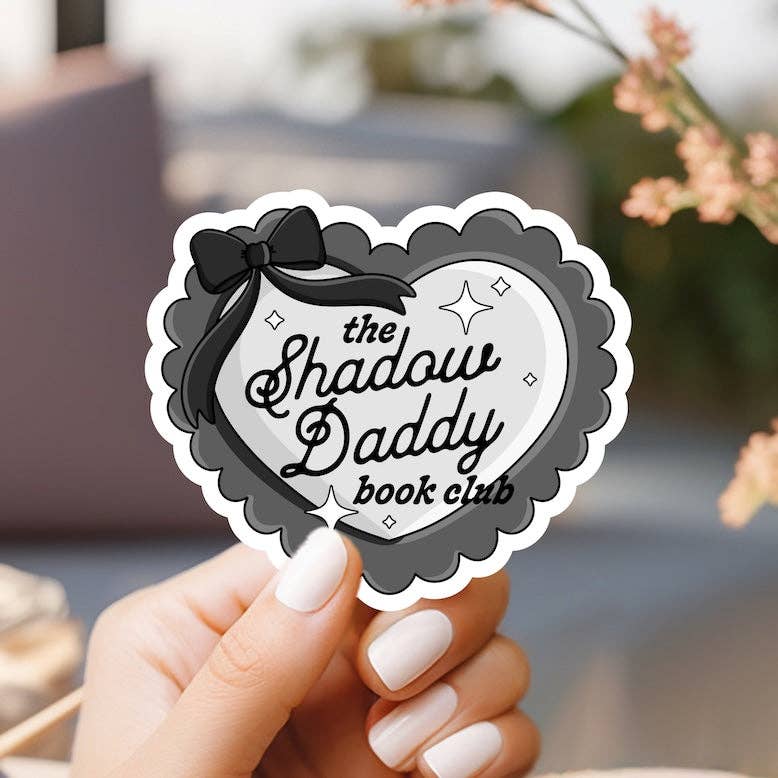 Heart-shaped sticker with 'the Shadow Daddy book club' text held by a hand with white nail polish.