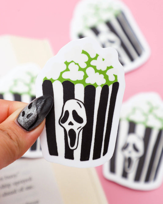 Popcorn cupcake sticker with ghost face design held by a hand on a pink background