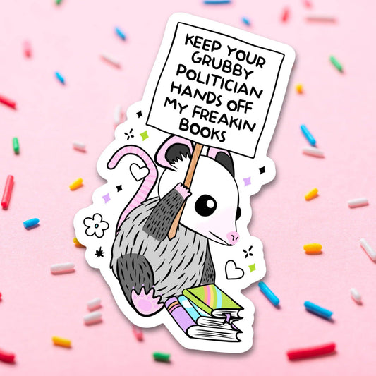 Sticker of an opossum holding a sign with text on a pink background with colorful sprinkles.