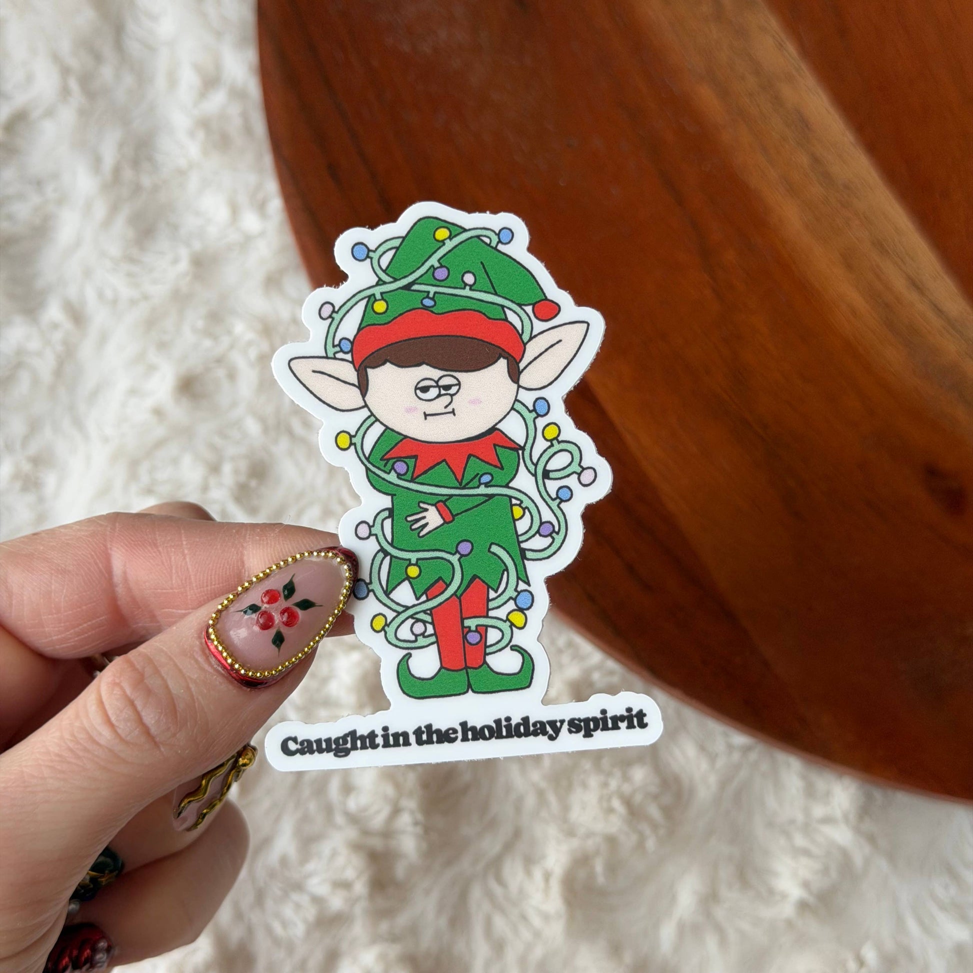 A hand holds a die-cut sticker featuring a cartoon elf wrapped in Christmas lights, with the text 'Caught in the holiday spirit' at the bottom. The background shows a white textured rug and a wooden surface.