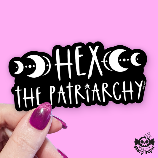 A hand with glittery purple fingernails holds a black die-cut sticker featuring white text reading 'HEX THE PATRIARCHY' with moon phases on either side, against a solid pink background. A small 'sharp sugar' logo appears in the bottom right corner.