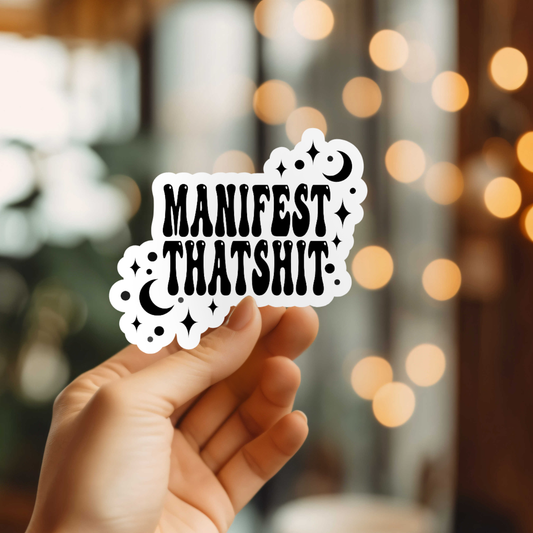 Hand holding a sticker with 'MANIFEST THATSHIT' text against a blurred bokeh background