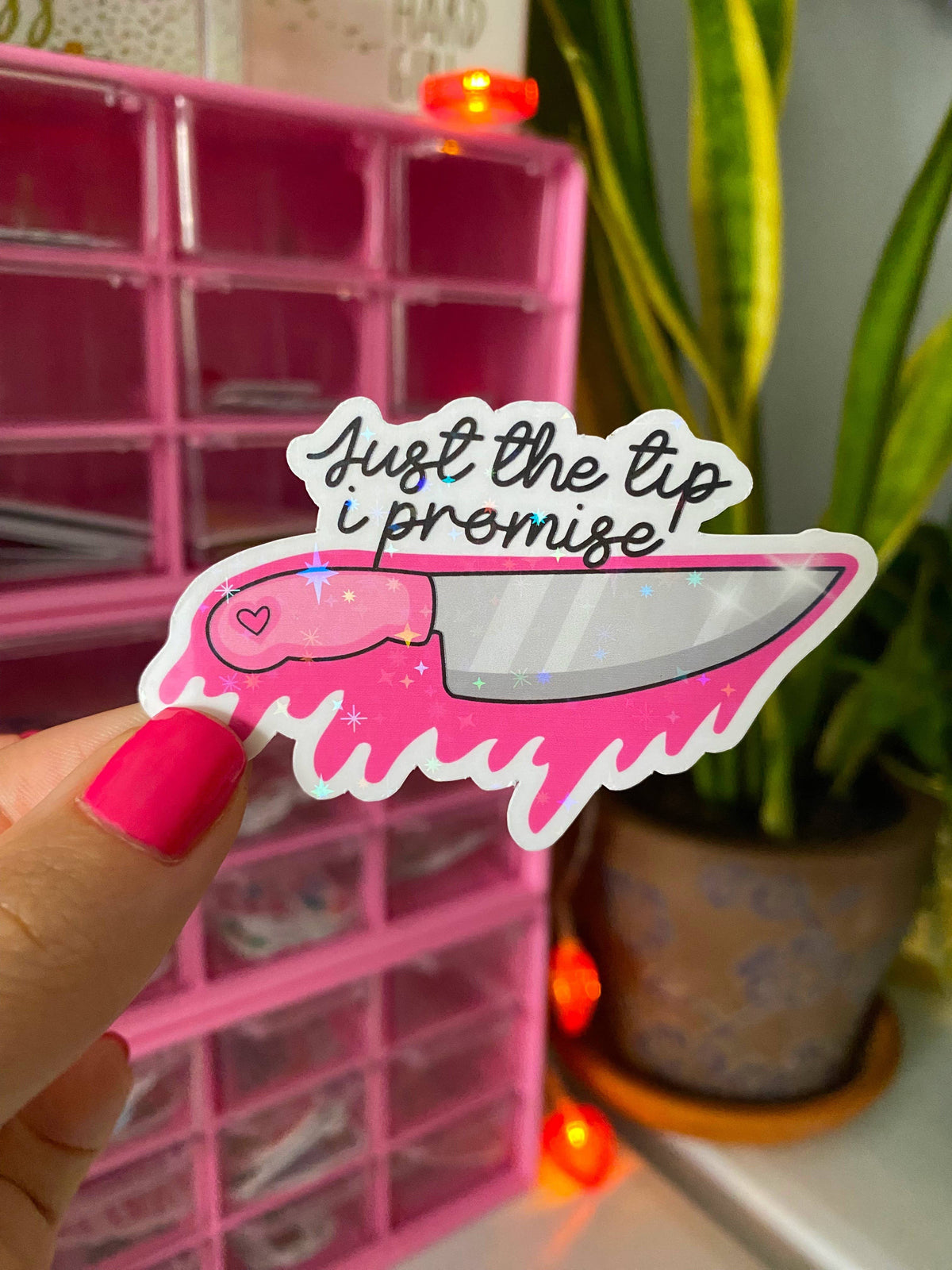 Sticker with knife design and text held by a hand in front of a pink organizer and plant.