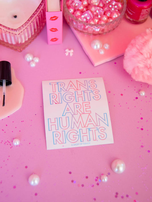 Card with 'Trans Rights are Human Rights' on a pink surface with decorative items.