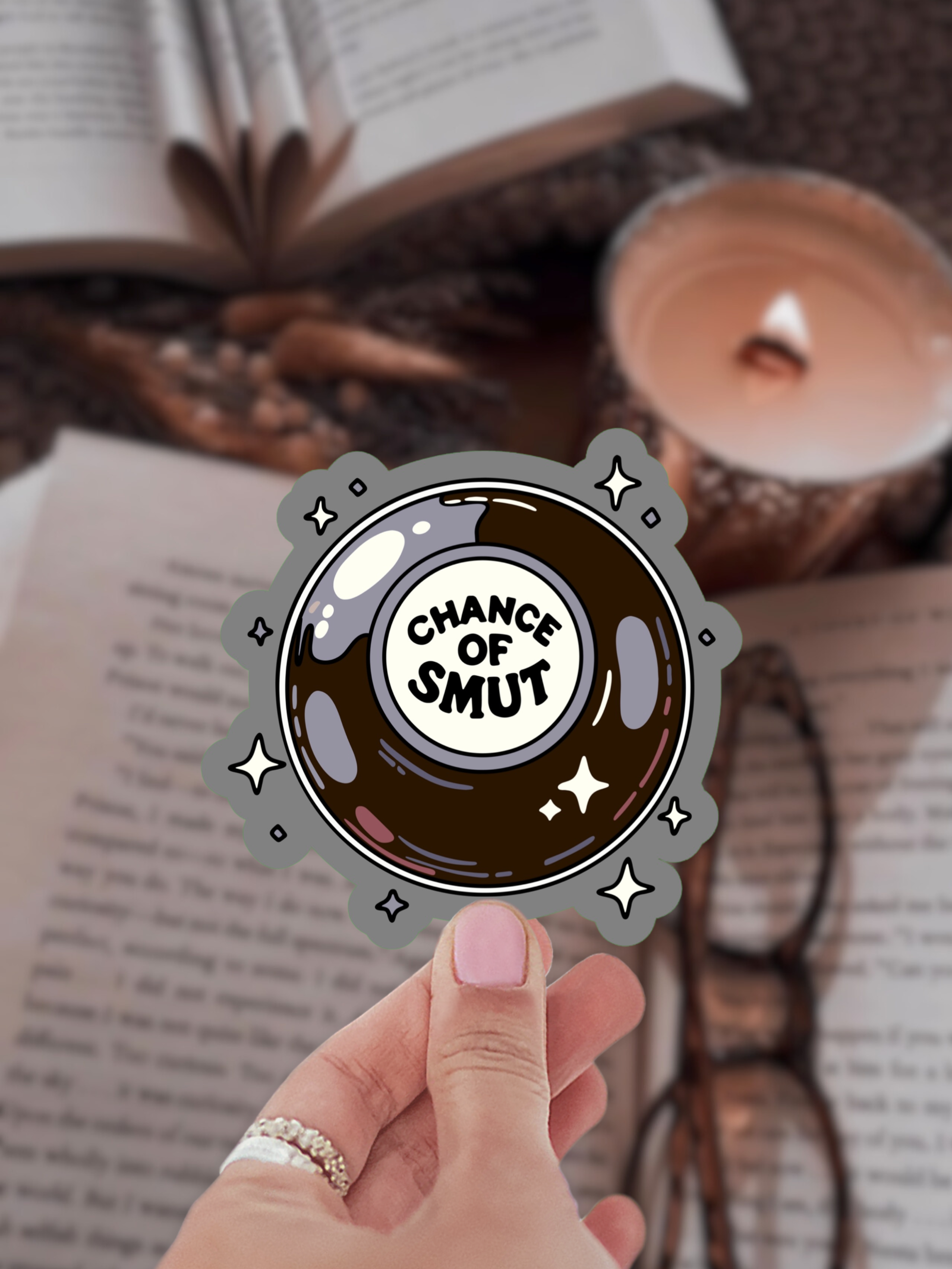 Hand holding a 'Chance of Smut' sticker over an open book with a candle in the background
