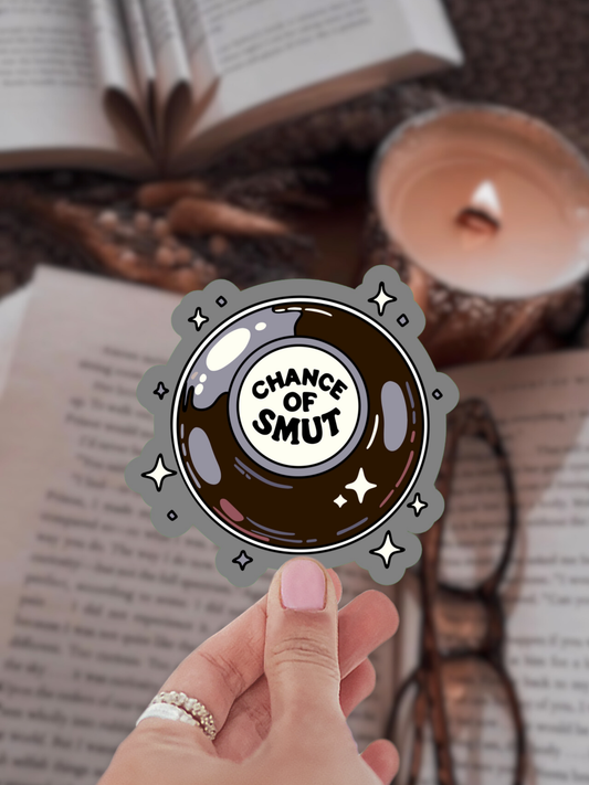 Hand holding a 'Chance of Smut' sticker over an open book with a candle in the background
