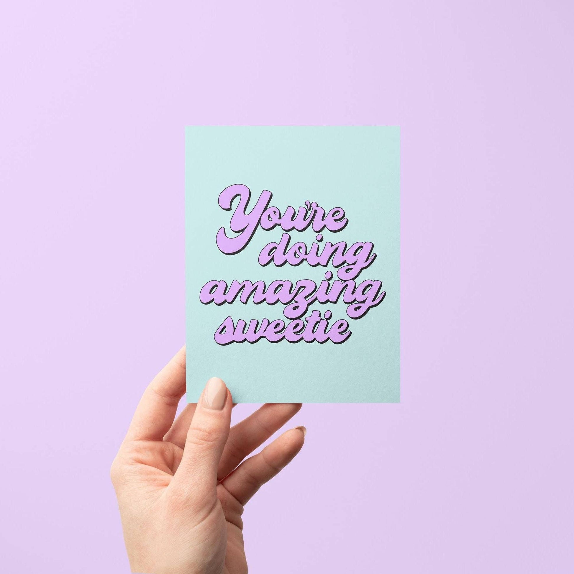 Hand holding a card with 'You're doing amazing sweetie' text on a pastel purple background