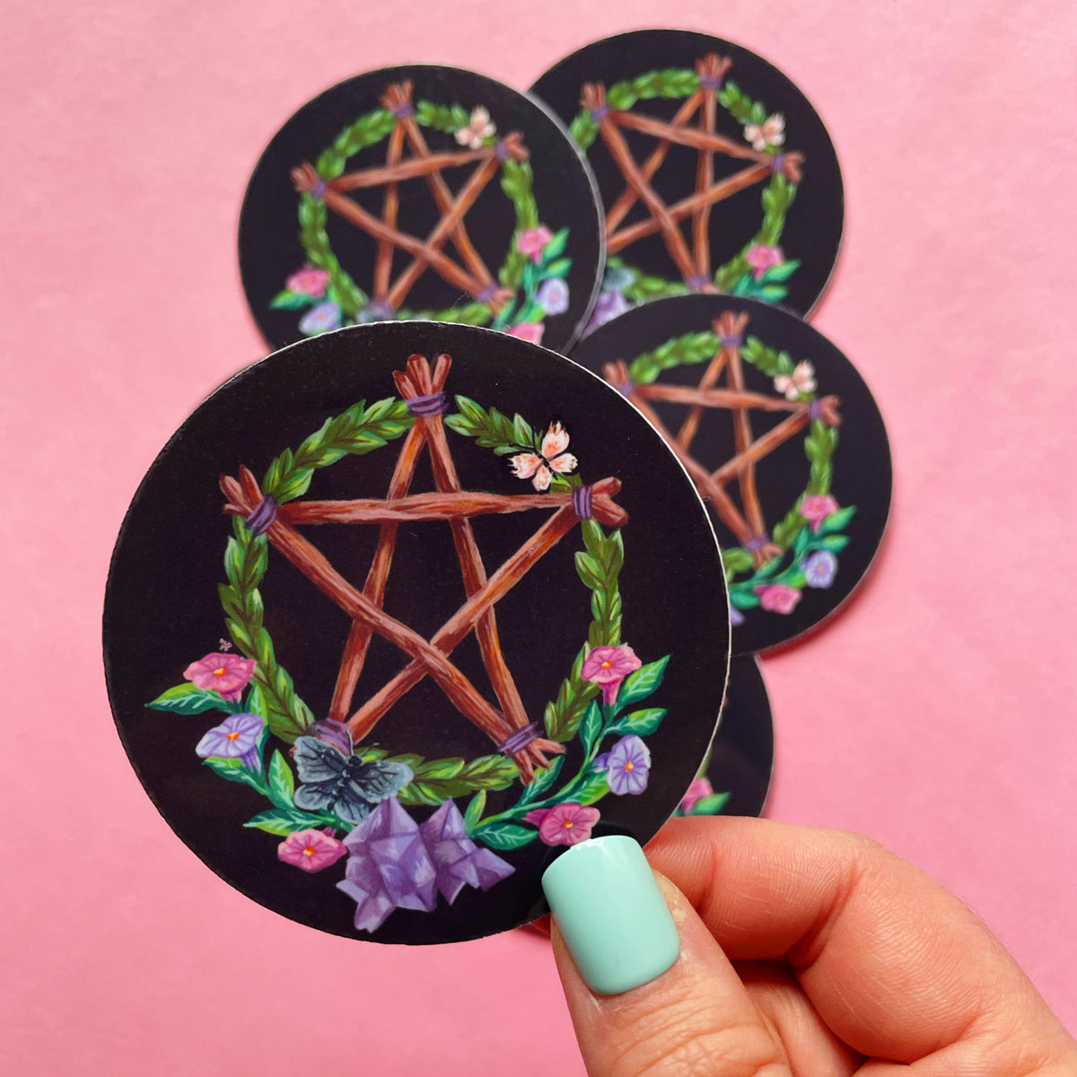 A hand with mint green nail polish holds a round black sticker featuring a wooden pentagram inside a green leaf wreath with pink and purple flowers. Three more identical stickers are stacked in the background against a solid pink backdrop.