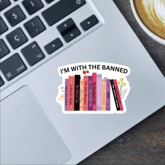 Laptop with a 'I'm with the banned' sticker featuring book spines on a desk with a cup of coffee.