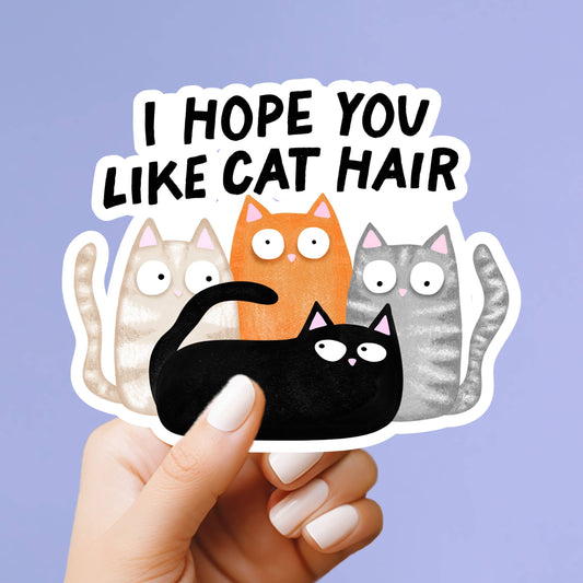 A hand with white manicured nails holds a die-cut sticker that reads 'I HOPE YOU LIKE CAT HAIR' above four cartoon cats in beige, orange, gray, and black, against a light lavender background.