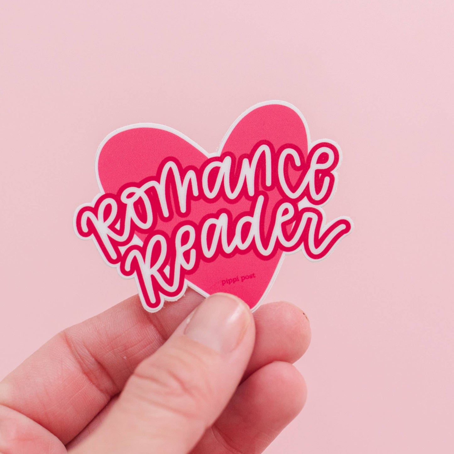 Heart-shaped sticker with 'Romance Reader' text held by a hand against a pink background