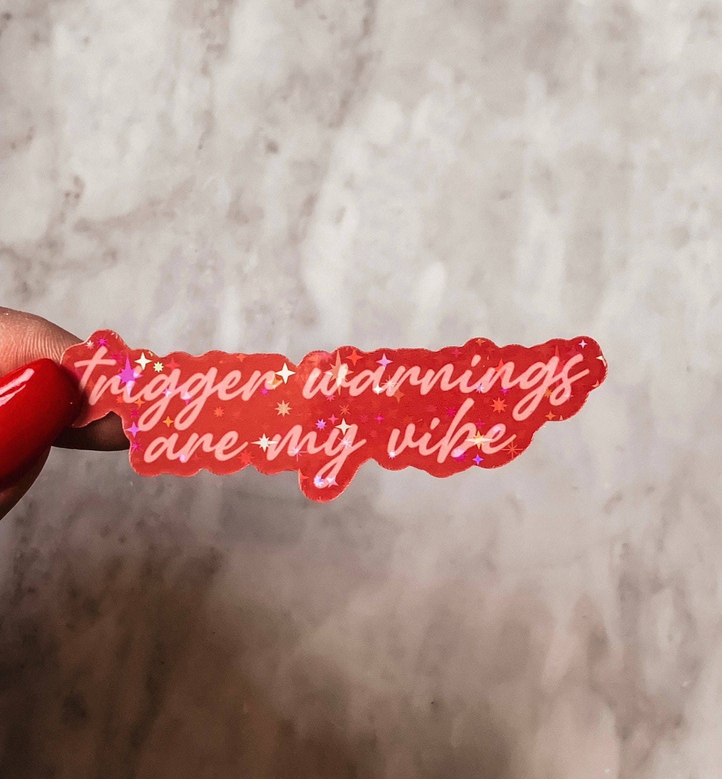 Red sticker with text 'trigger warnings are my vibe' held against a marble background