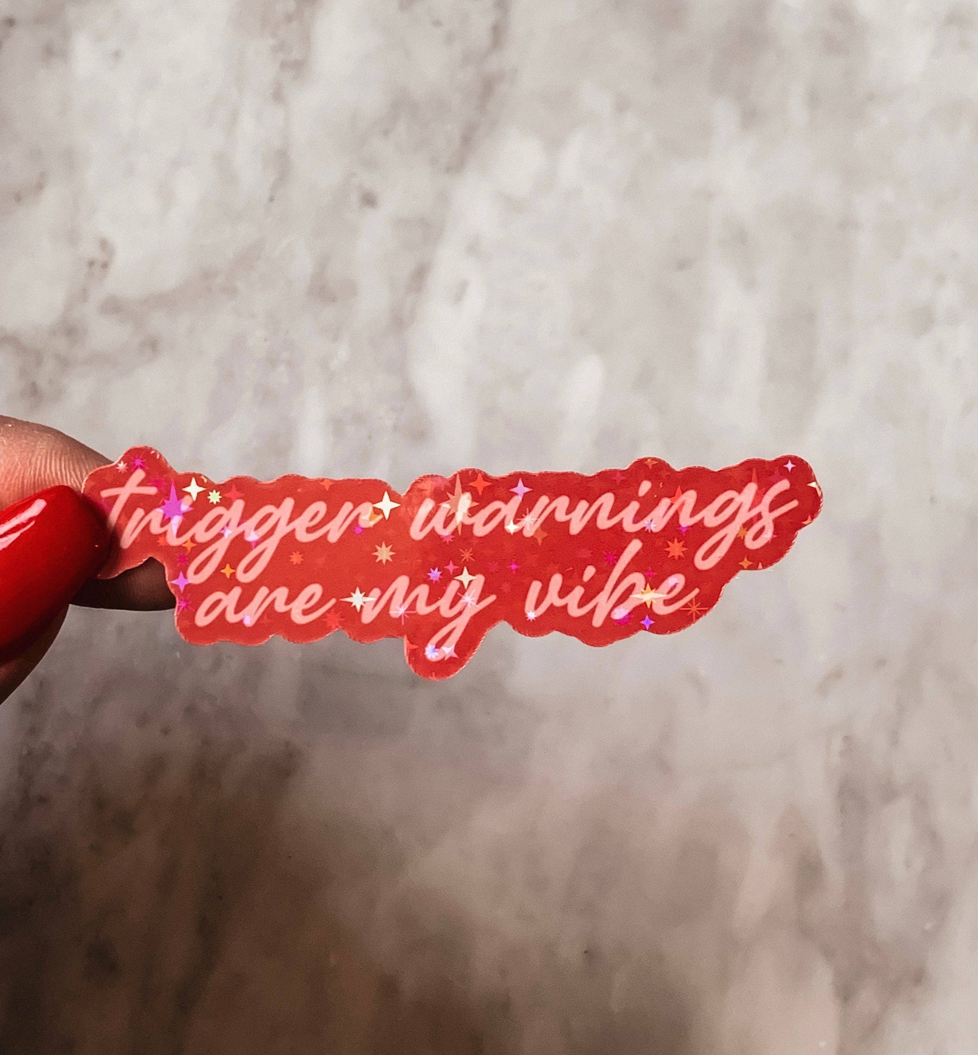 Red sticker with text 'trigger warnings are my vibe' held against a marble background
