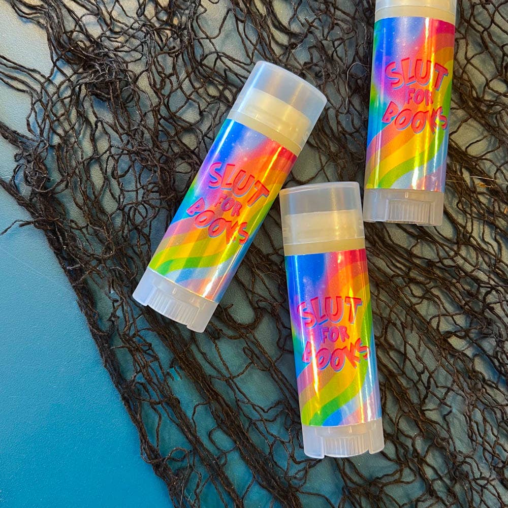 Three rainbow-colored lip balms with 'SLUT FOR LUNCH' text on a textured surface.