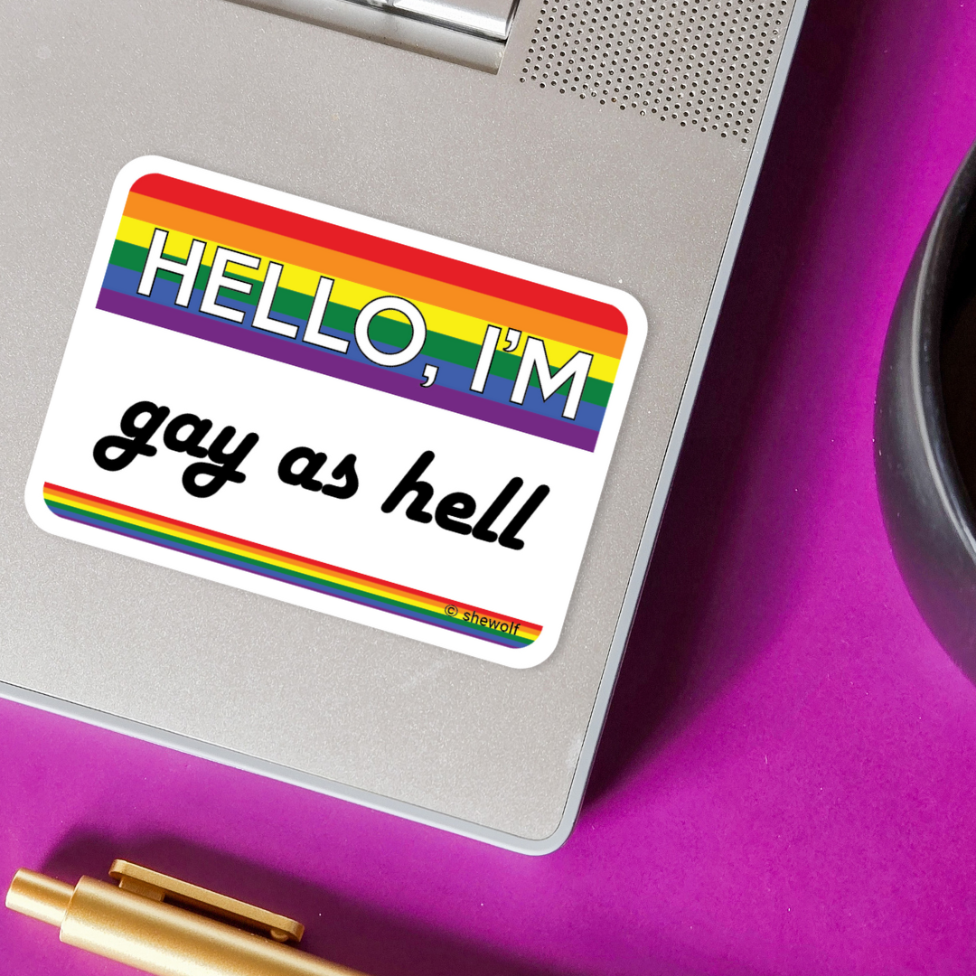 Sticker with rainbow border and text 'HELLO, I'M gay as hell' on a metallic surface.
