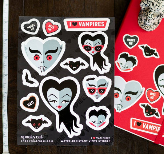 A sheet of vampire-themed water-resistant vinyl stickers on a dark wood background, next to a red sticker album cover. The stickers feature cartoon vampire faces, bats, and heart-shaped sweets with text like 'HAUNT ME' and 'BITE ME'.