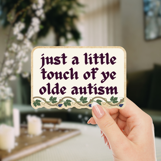 Hand holding a sign with text 'just a little touch of ye olde autism' against a blurred indoor background