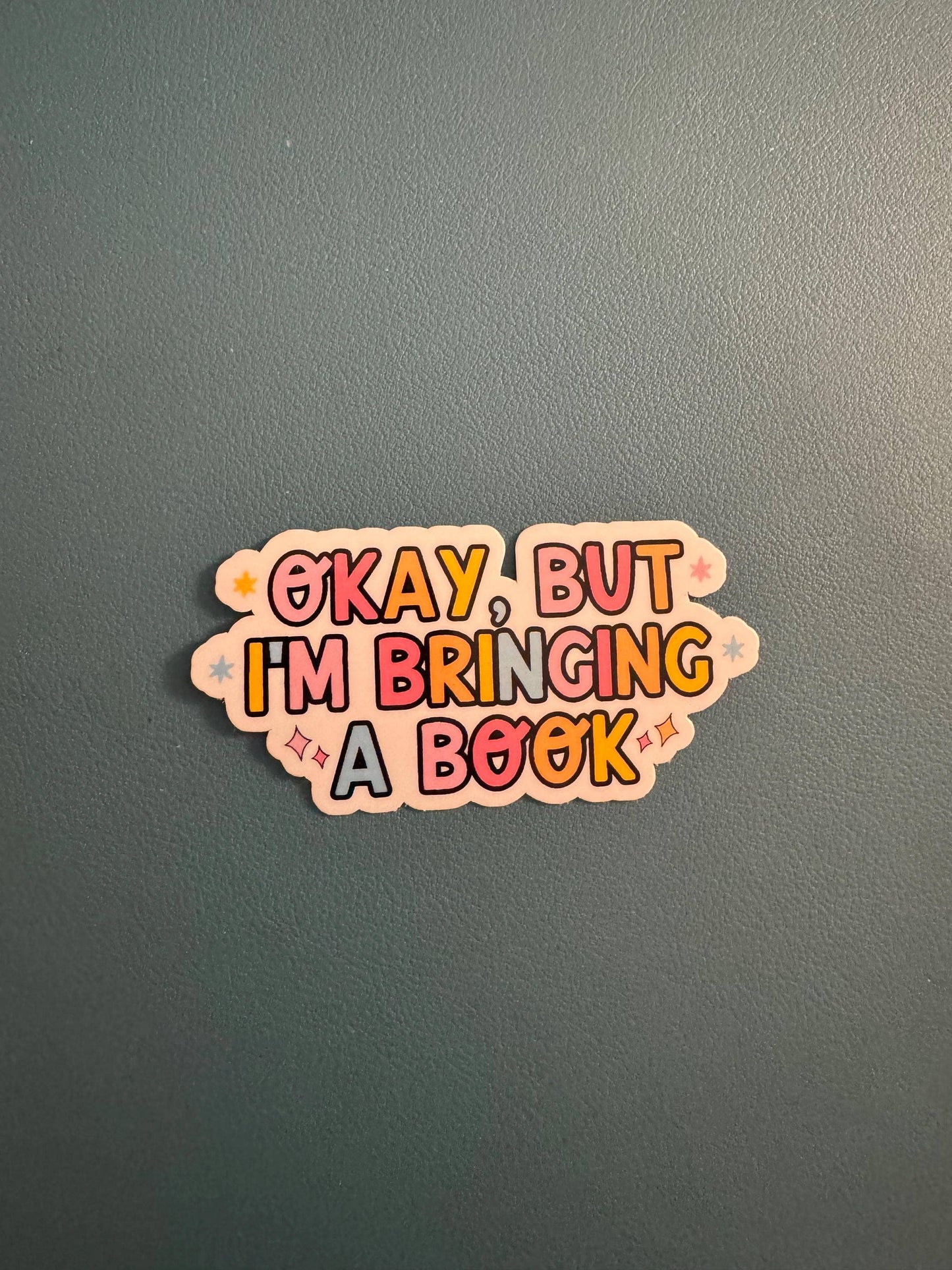 A die-cut sticker with the text 'OKAY, BUT I'M BRINGING A BOOK' in colorful bubble letters on a dark gray textured background.