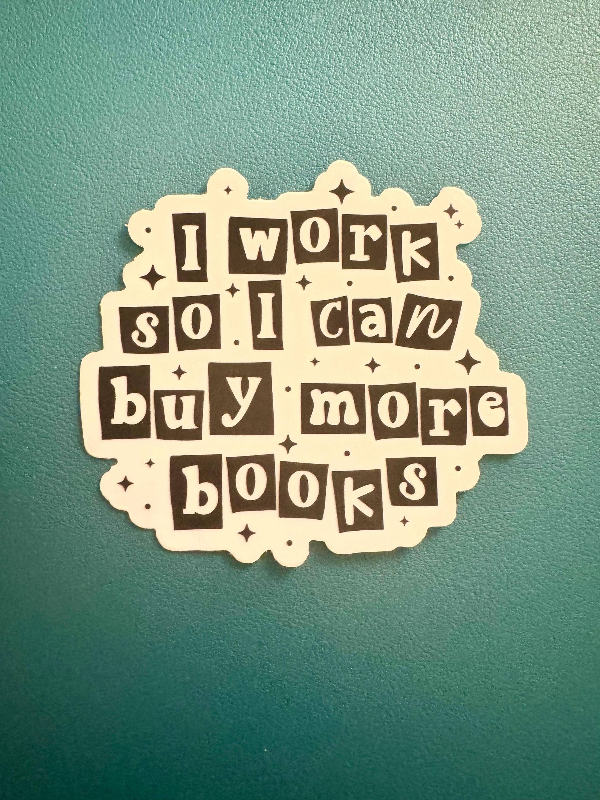 A die-cut sticker with the phrase 'I work so I can buy more books' in white letters on dark brown blocks, placed on a textured teal-green background.