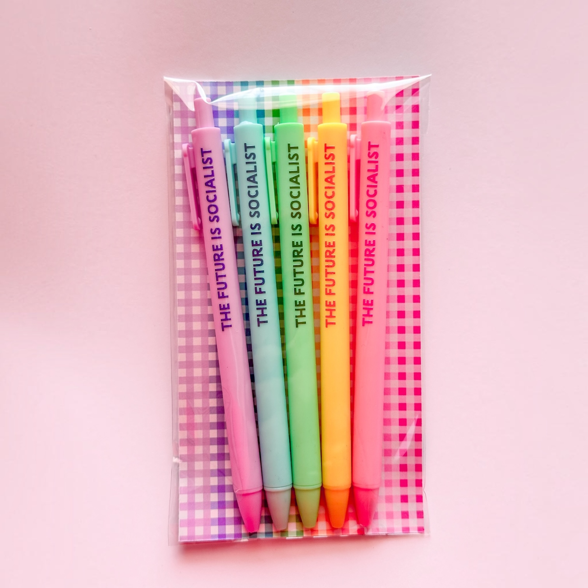 A set of six pastel-colored pens in a clear plastic sleeve with a pink and white gingham-patterned backing card. Each pen is printed with the text 'THE FUTURE IS SOCIALIST' in dark purple.