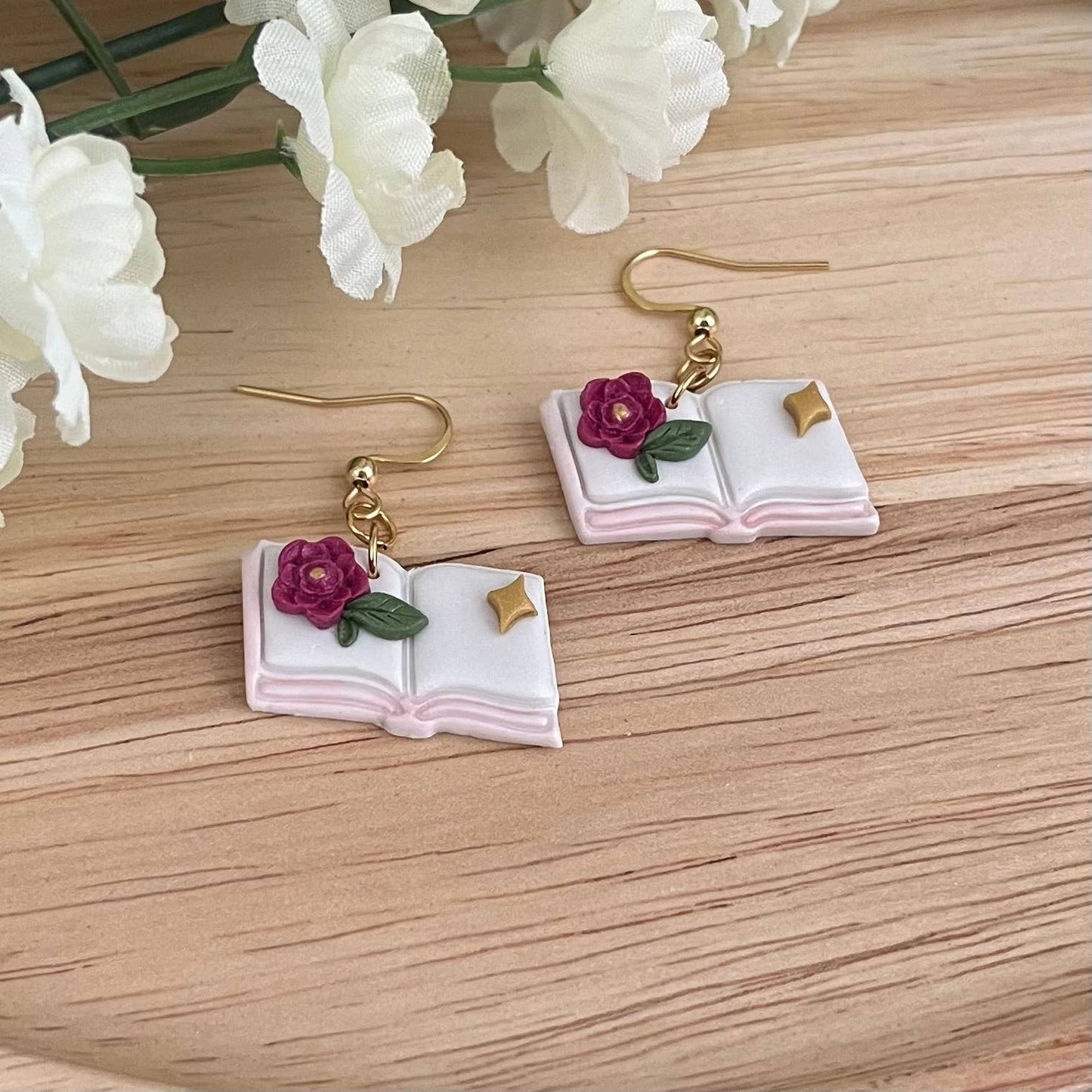 Book-shaped earrings with floral details on a wooden surface with white flowers.