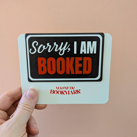 Hand holding a magnetic bookmark with 'Sorry, I am booked' text on a beige background