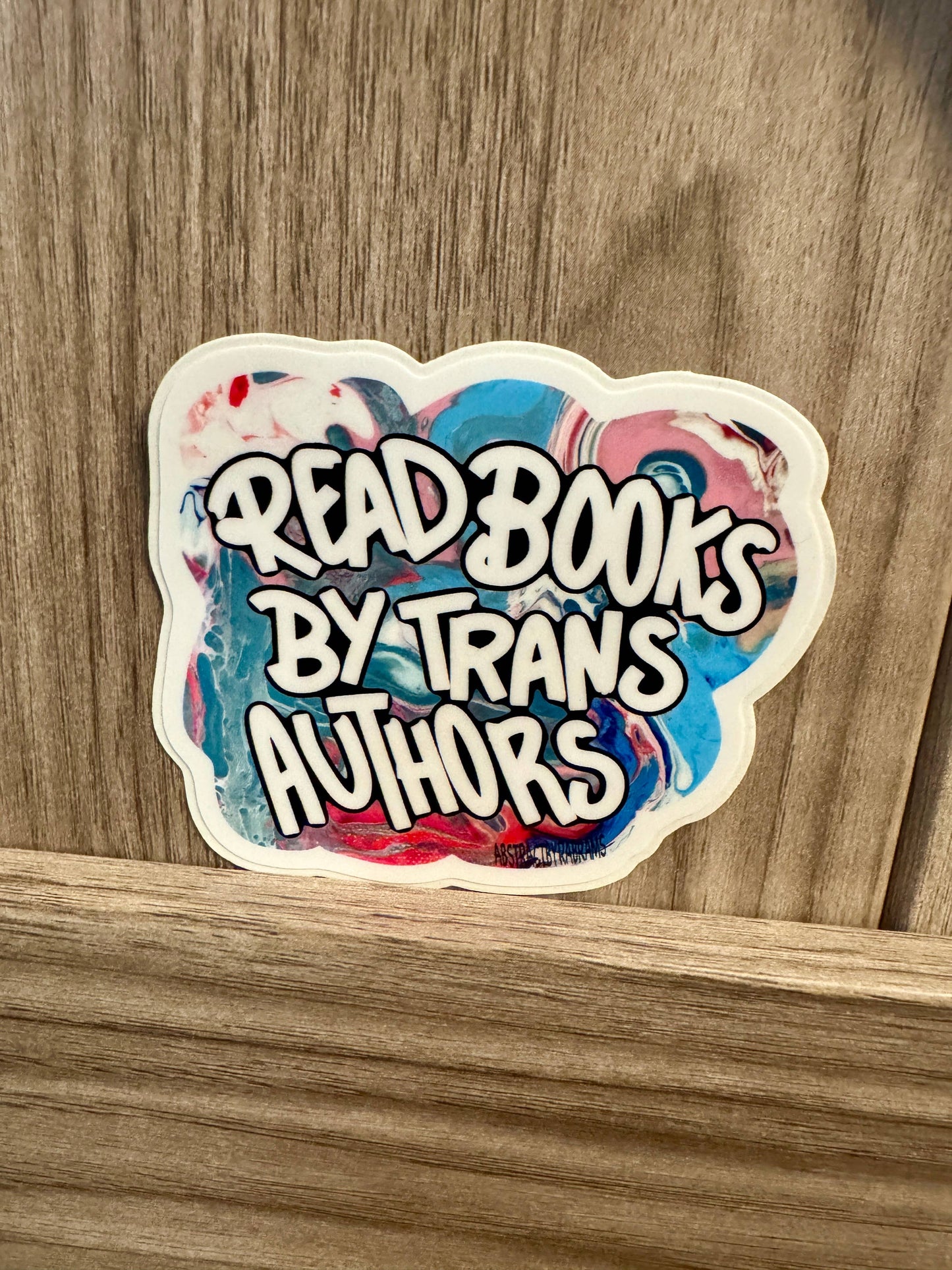 Sticker with 'Read Books by Trans Authors' text on a wooden surface