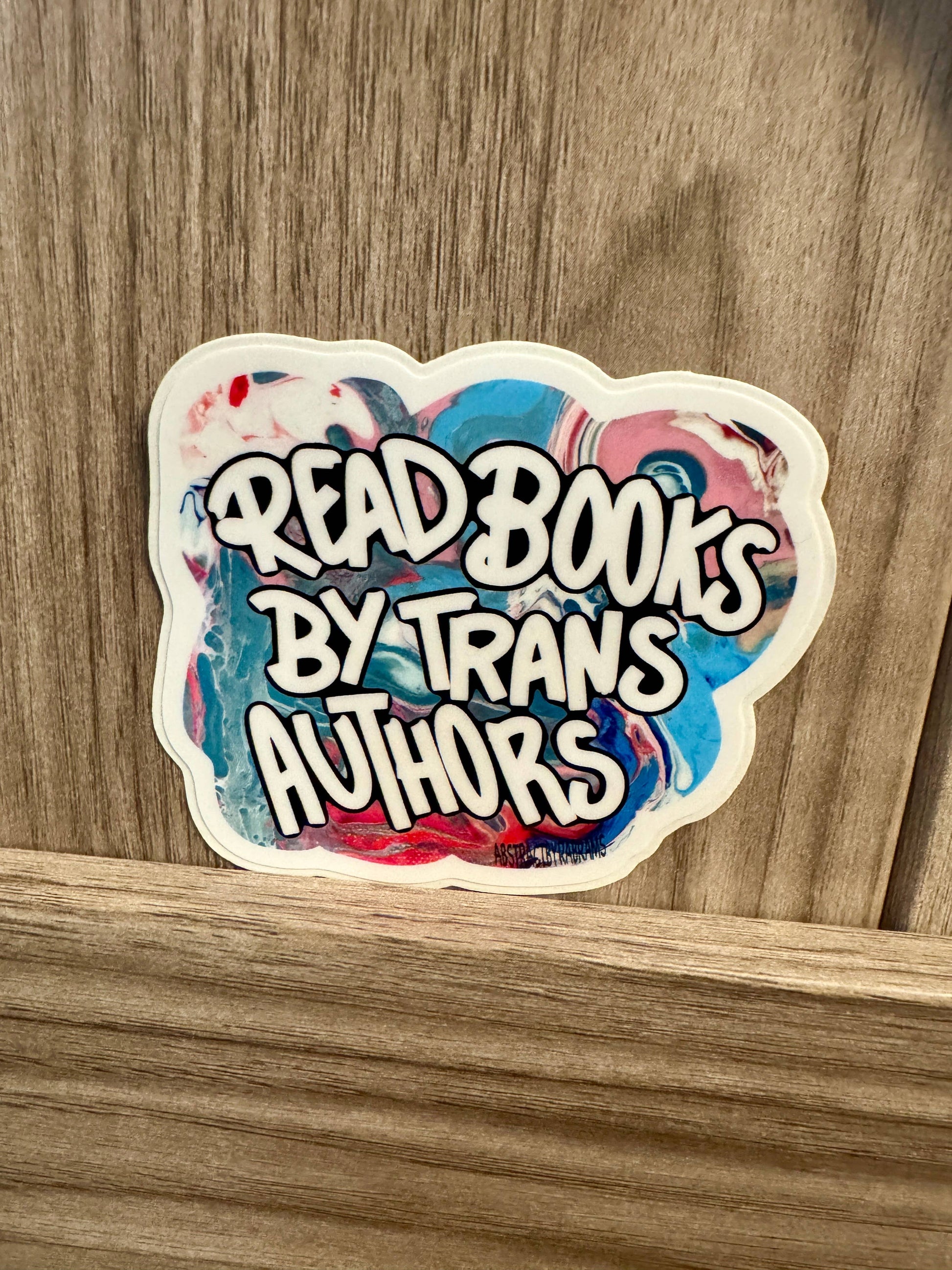 Sticker with 'Read Books by Trans Authors' text on a wooden surface