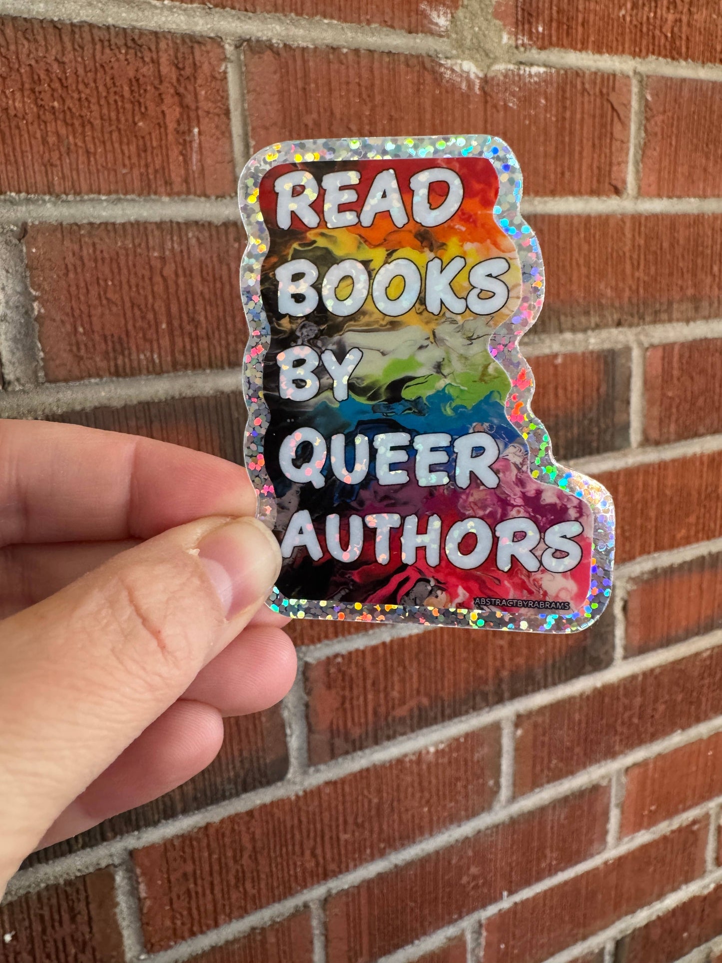 Holographic sticker with text 'Read books by queer authors' held against a brick wall.