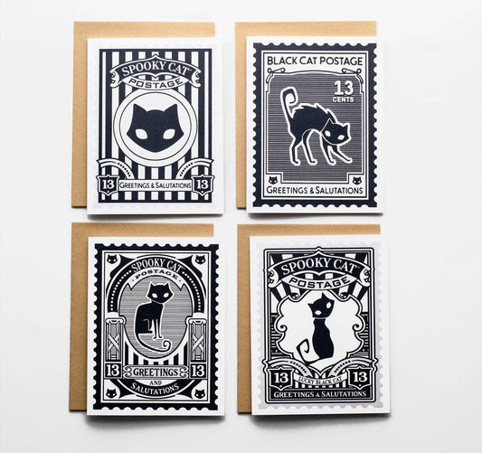 Four Halloween-themed postage stamp greeting cards arranged in a 2x2 grid on a white background, each with a kraft envelope behind it. The stamps feature black cats, vintage typography, and designs in black, white, and navy tones.