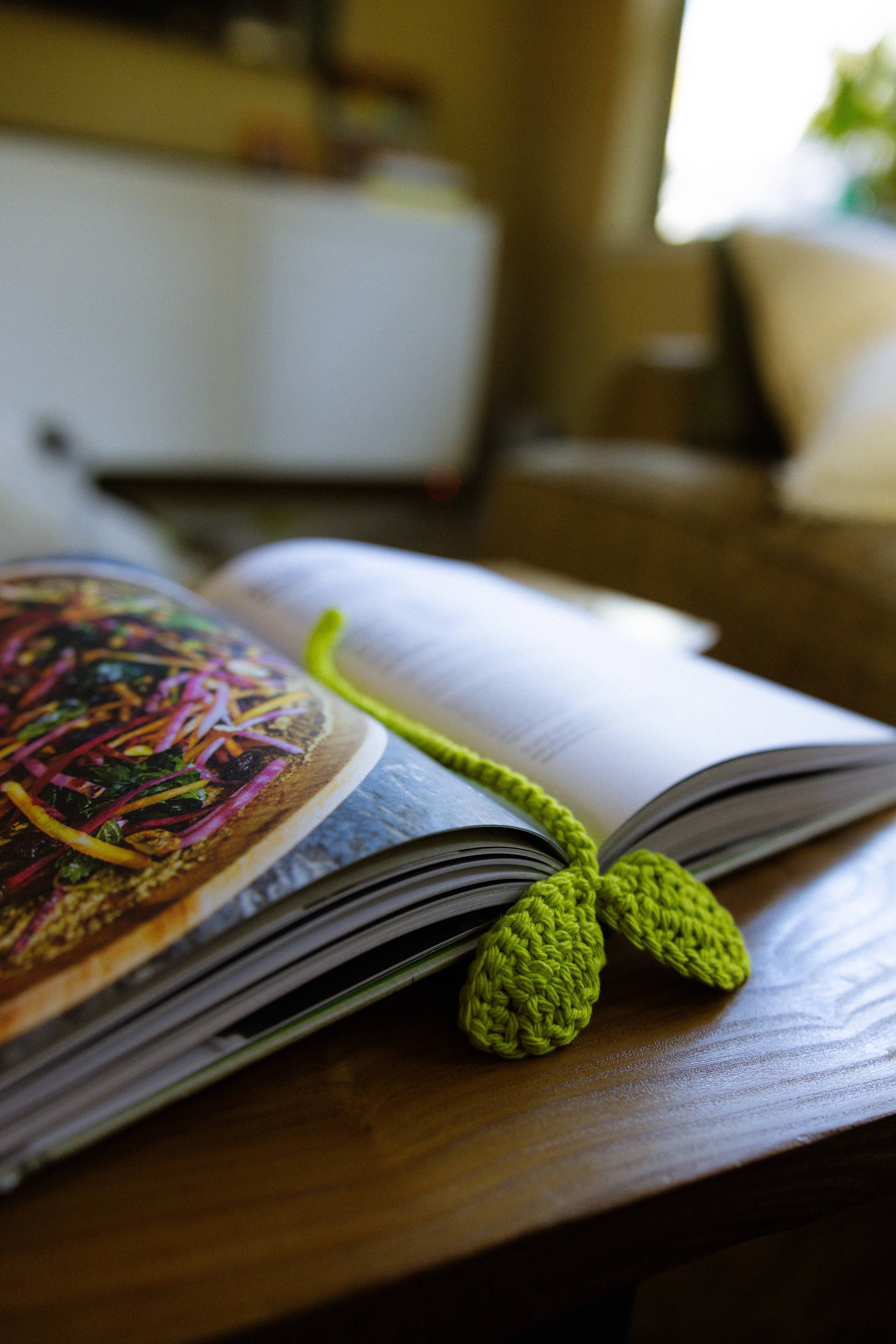 Open book with a green knitted bookmark on a wooden surface, blurred indoor background