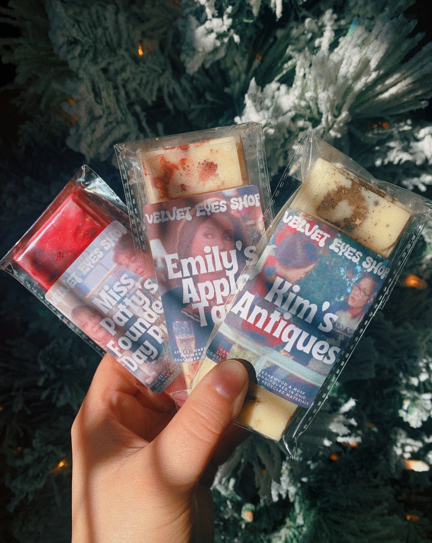 Hand holding three wax melts with visible brand names against a Christmas tree background