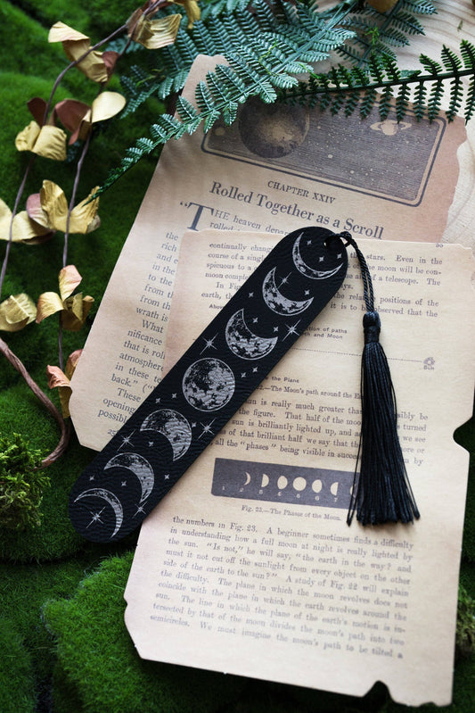 Black leather bookmark with moon phases and tassel on an open book with greenery.