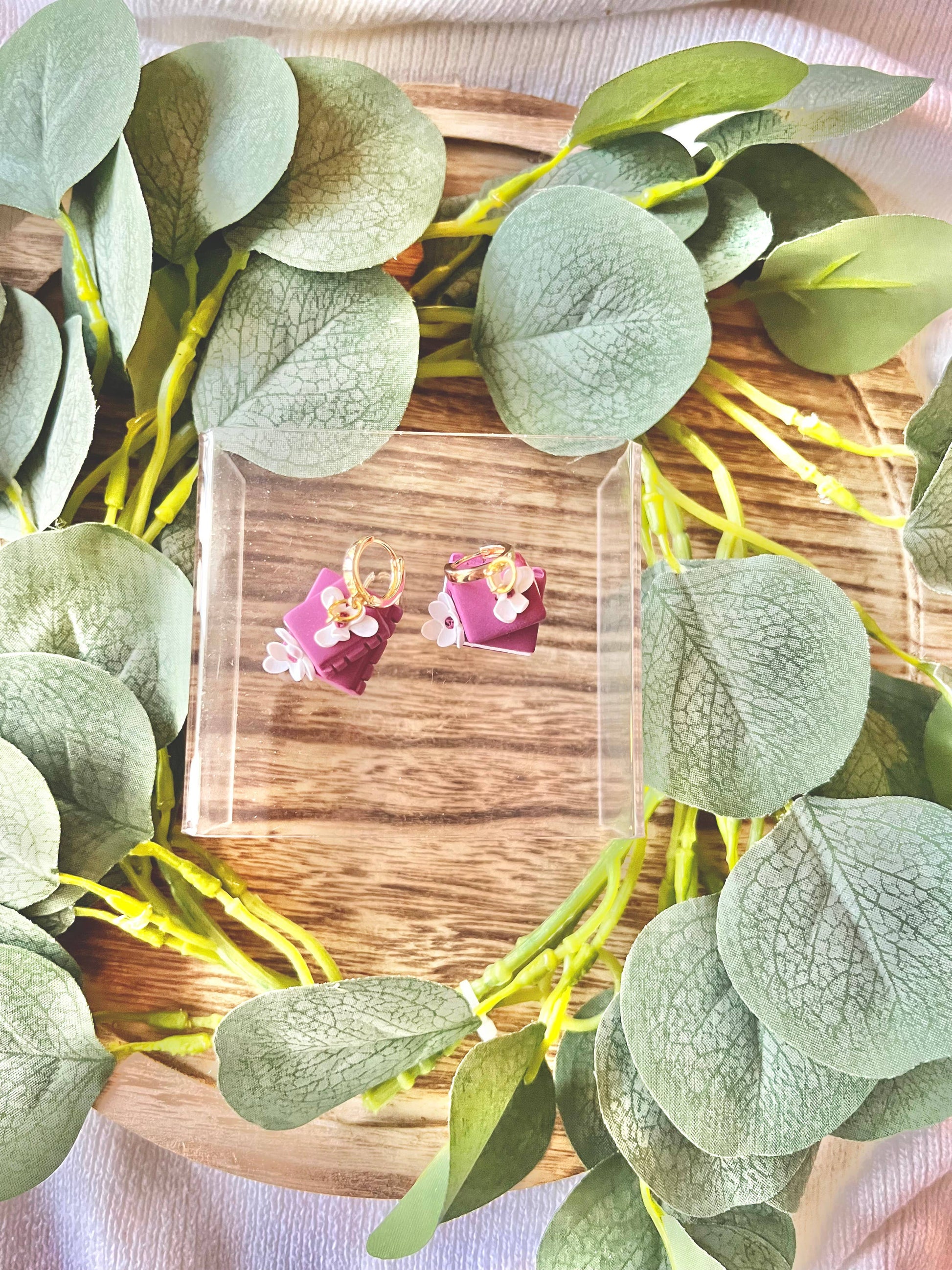 Pair of pink earrings with floral design on a wooden board surrounded by green leaves.