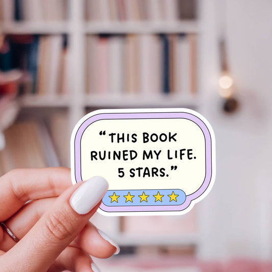 Sticker with text 'This book ruined my life. 5 stars.' held by a hand in front of a blurred bookshelf.