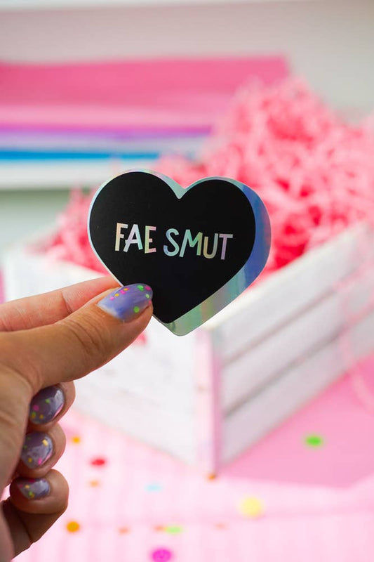 A hand with glittery purple nail polish holds a holographic heart-shaped sticker that reads 'FAE SMUT' in white letters. The background is a soft-focus pink and white setting with decorative confetti.
