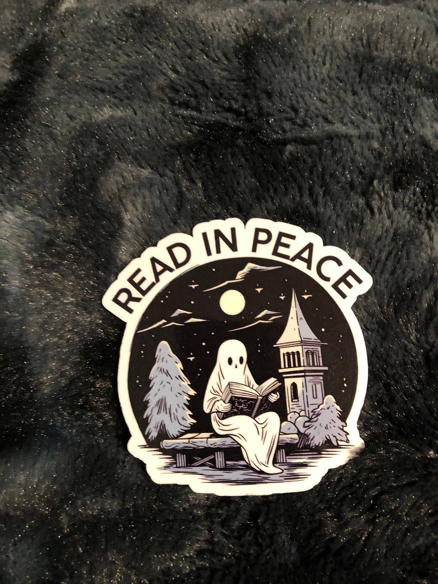 A die-cut sticker featuring a ghost reading a book under a full moon near a church tower, with the words 'READ IN PEACE' arched above, placed on a black furry surface.
