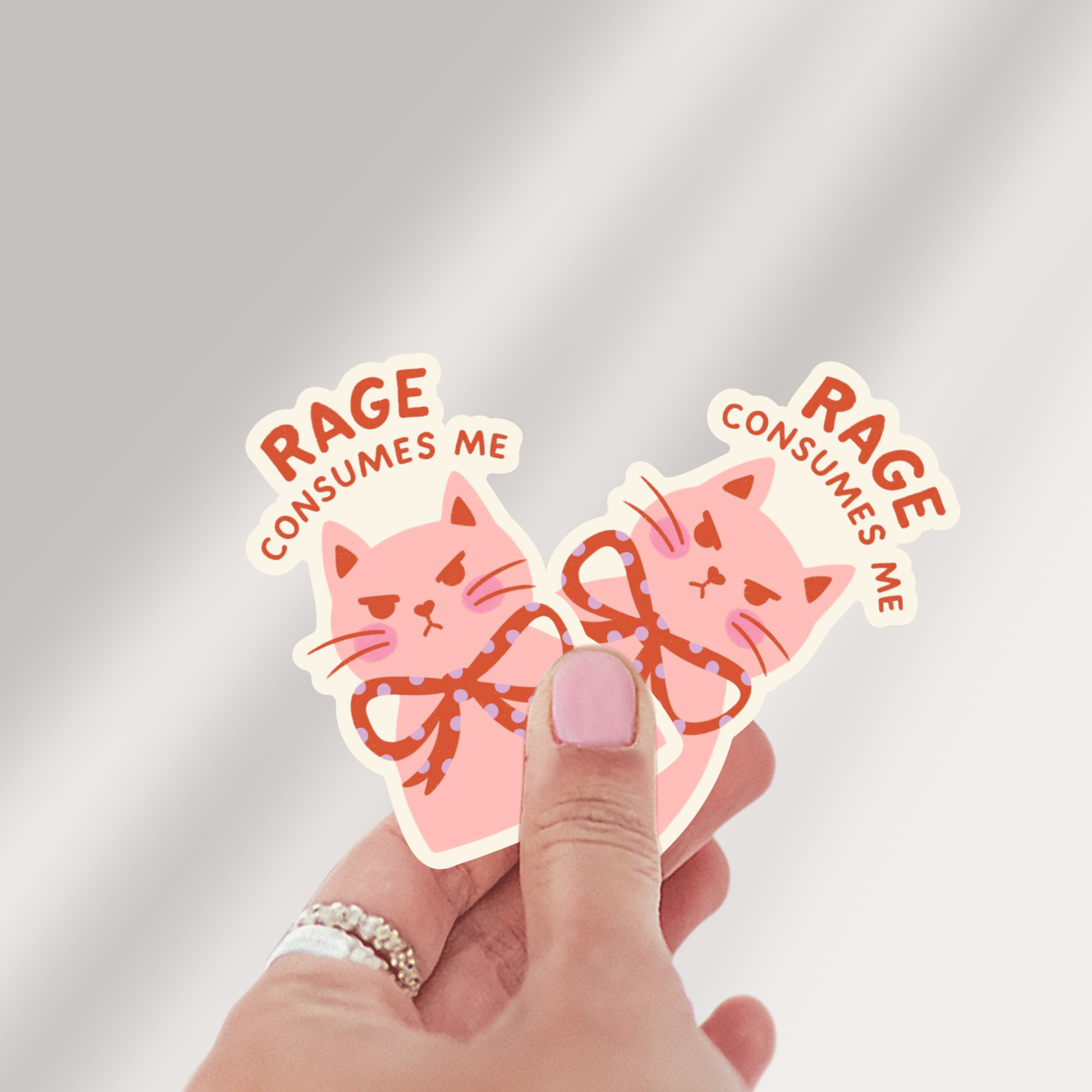Two pink cat-shaped stickers with 'RAGE CONSUMES ME' text held by a hand against a light gray background.
