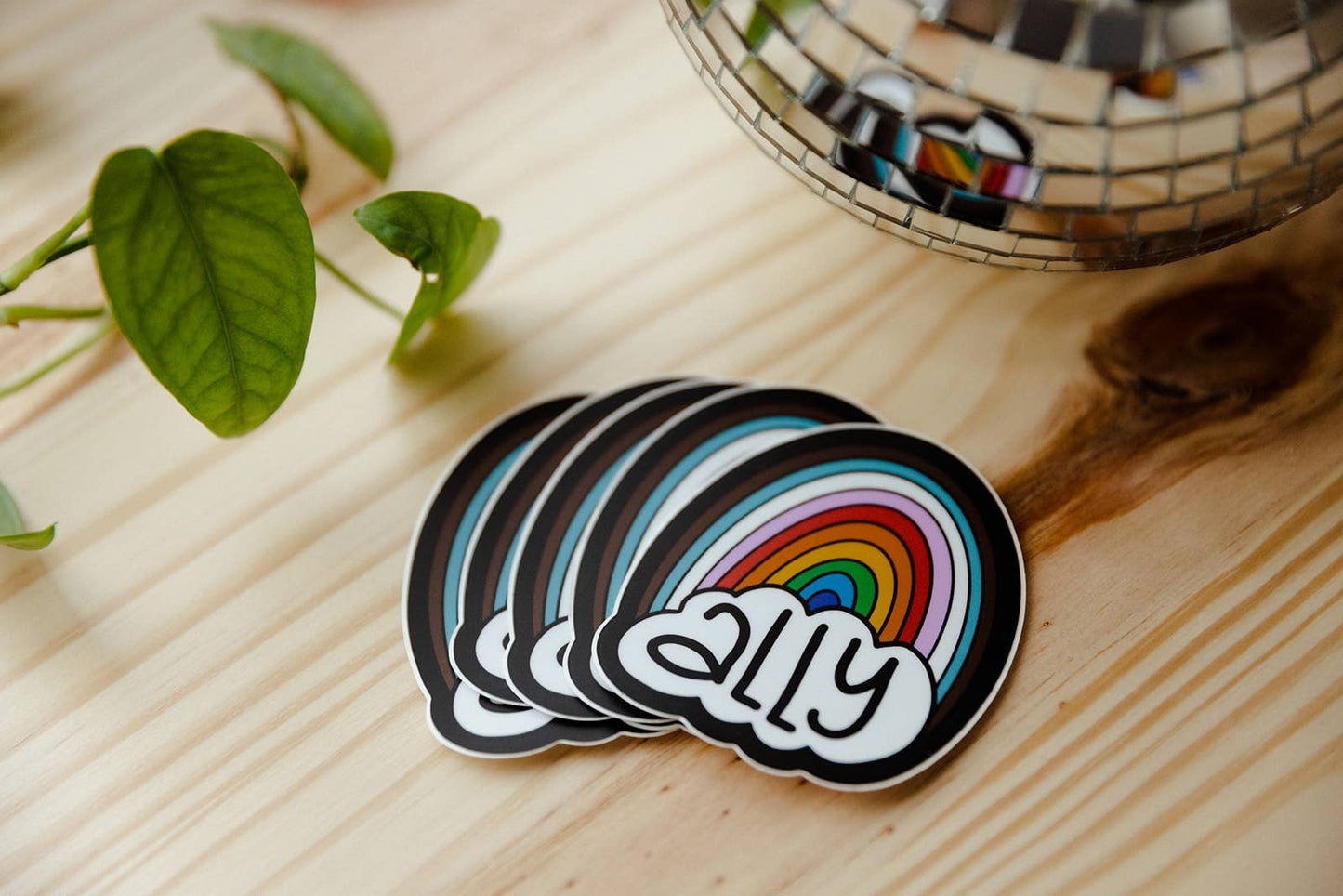 Stack of stickers with rainbow design and 'ally' text on a wooden surface with a plant and disco ball in the background.