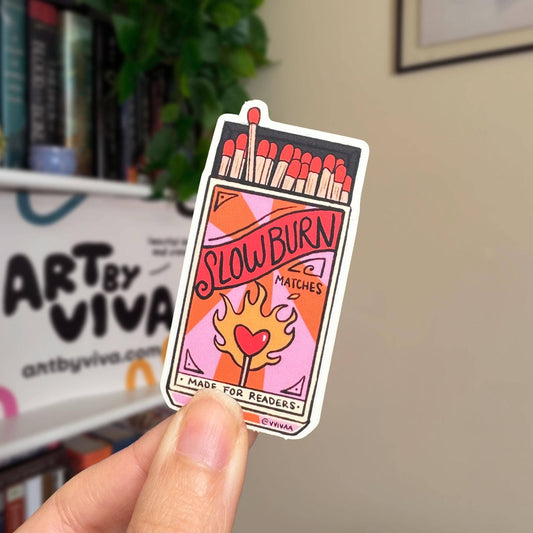 Sticker of a matchbox with 'Slow Burn' text held by a hand, with a blurred indoor background.