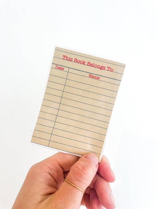 Hand holding a small piece of paper with 'This Book Belongs To' text on a white background