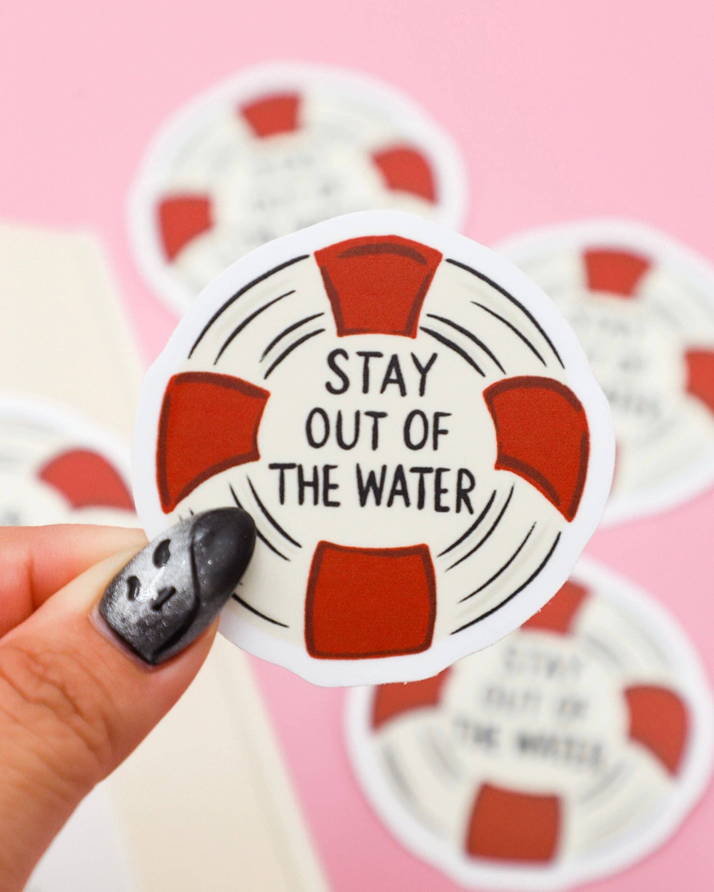 Life buoy-shaped sticker with 'Stay out of the water' text held by a hand on a pink background