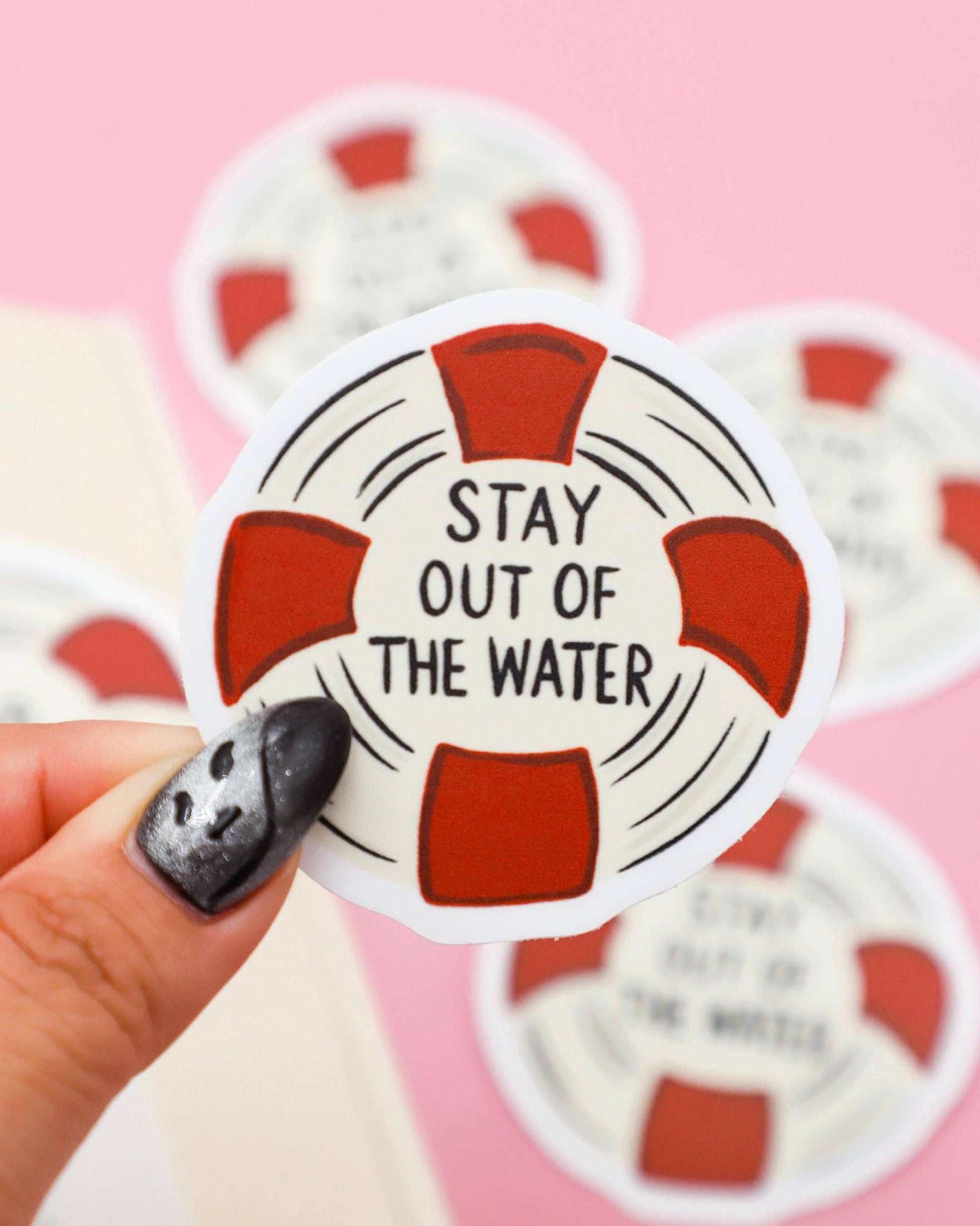 Life buoy-shaped sticker with 'Stay out of the water' text held by a hand on a pink background