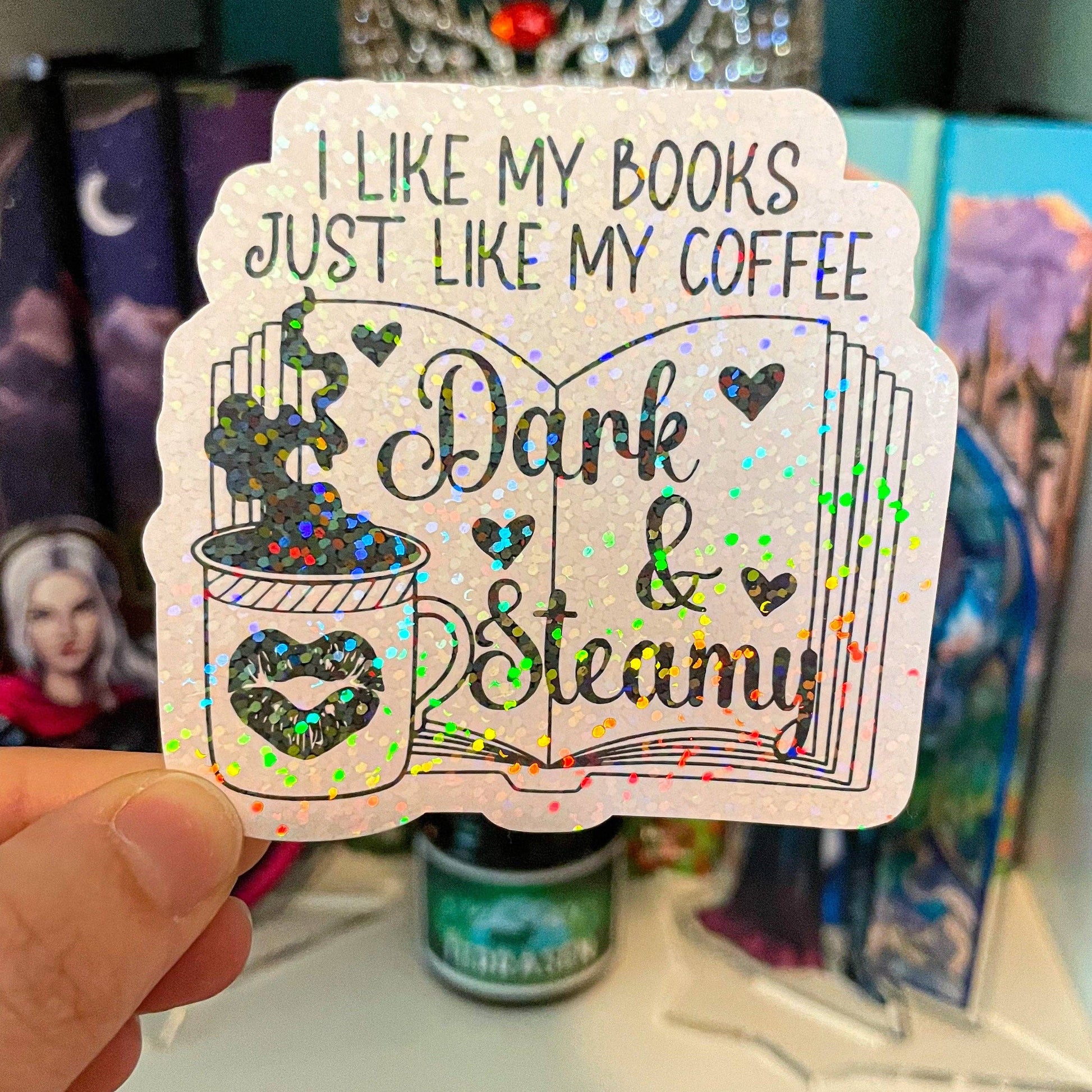 A hand holds a die-cut sticker featuring a glittery white background with the text 'I LIKE MY BOOKS JUST LIKE MY COFFEE' and an illustration of an open book and coffee mug decorated with multicolor holographic glitter.