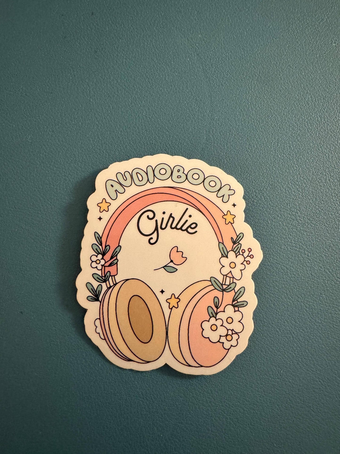 A die-cut sticker featuring a retro-style illustration with the words 'AUDIOBOOK Girlie' under a pink and orange rainbow, decorated with headphones, flowers, and stars, placed on a textured dark teal surface.