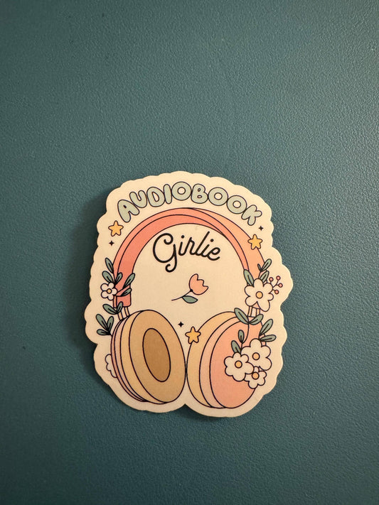 A die-cut sticker featuring a retro-style illustration with the words 'AUDIOBOOK Girlie' under a pink and orange rainbow, decorated with headphones, flowers, and stars, placed on a textured dark teal surface.