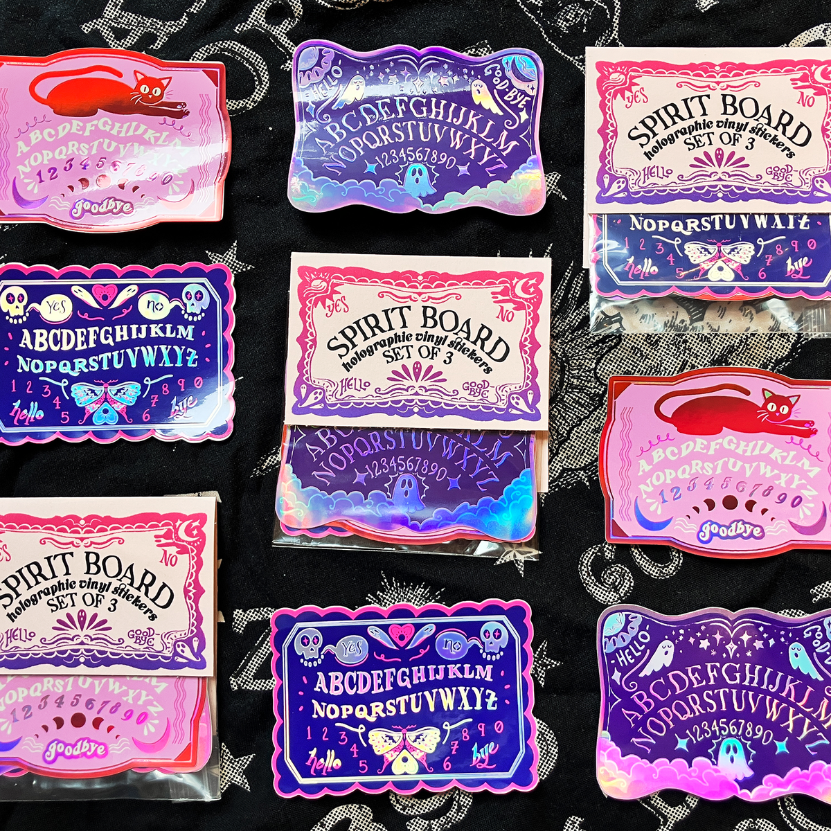 A set of colorful, holographic 'Spirit Board' and 'Hill's Logs' occult-themed sticker sheets arranged on a black fabric background with white spiderwebs.