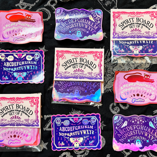 A set of colorful, holographic 'Spirit Board' and 'Hill's Logs' occult-themed sticker sheets arranged on a black fabric background with white spiderwebs.