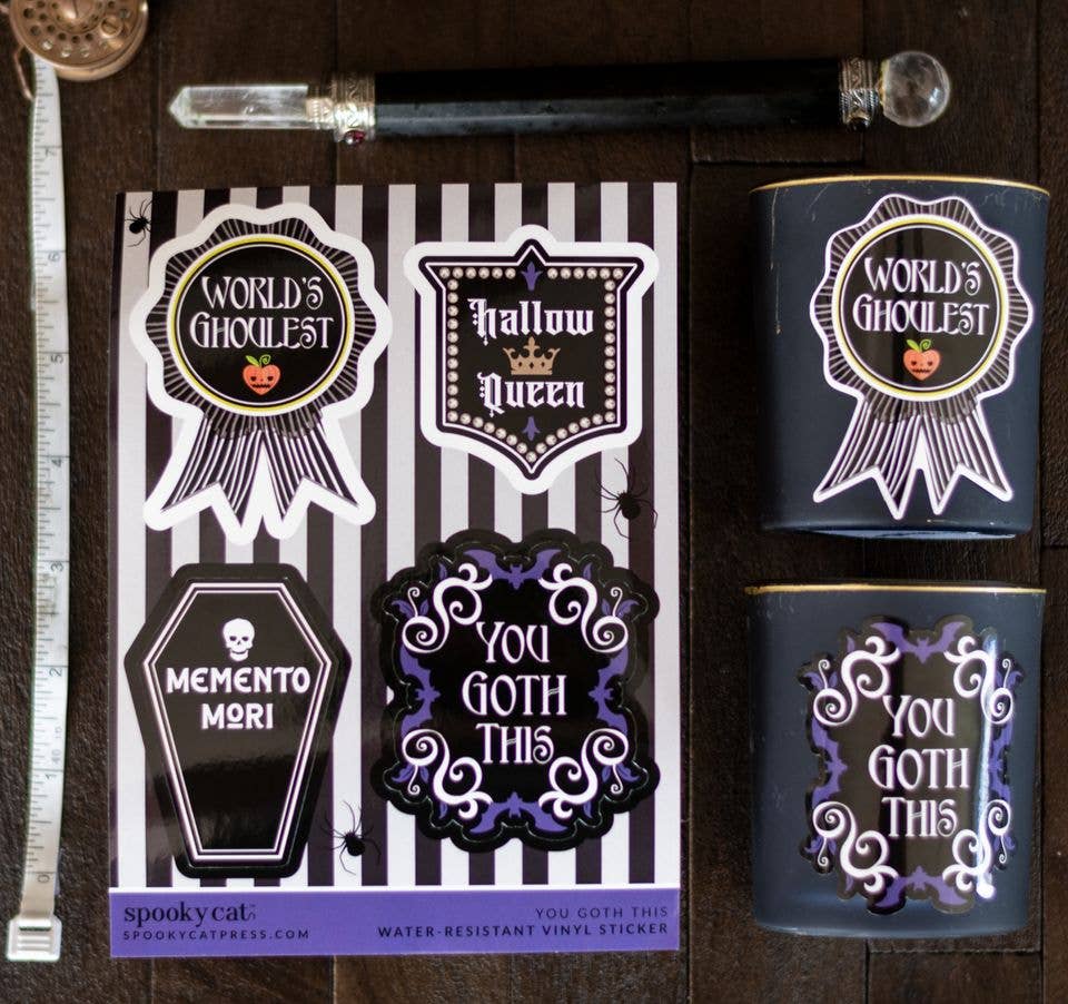 Halloween-themed vinyl stickers displayed on a dark wood background, including a sticker sheet with designs like 'World's Ghoulest', 'Halloween Queen', 'Memento Mori', and 'You Goth This', alongside two matching dark blue jars.