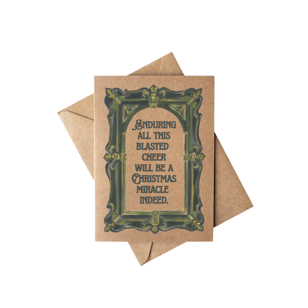 A square kraft paper greeting card with a decorative dark green ornate border and humorous text reading 'ENDURING ALL THIS BLASTED CHEER WILL BE A CHRISTMAS MIRACLE INDEED.' A matching kraft envelope is placed behind it on a clean white background.